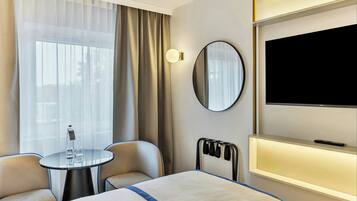 Ein Hotelzimmer mit Bett, Fernseher, Nachttisch mit Gläsern, Stuhl und einem Fenster mit Vorhängen.