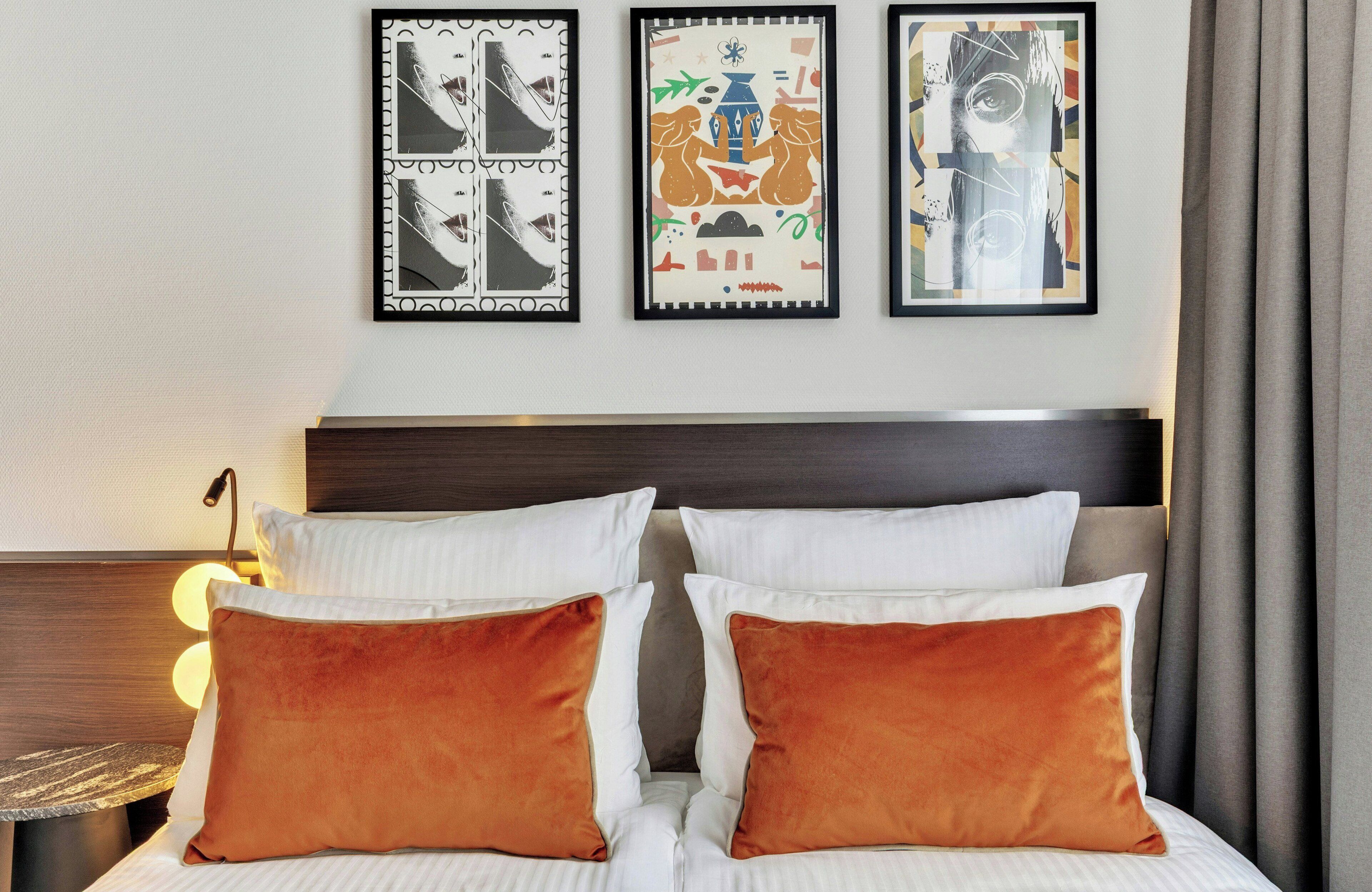 A hotel room with a bed, two pillows, a nightstand, a lamp, and three framed artworks on the wall.