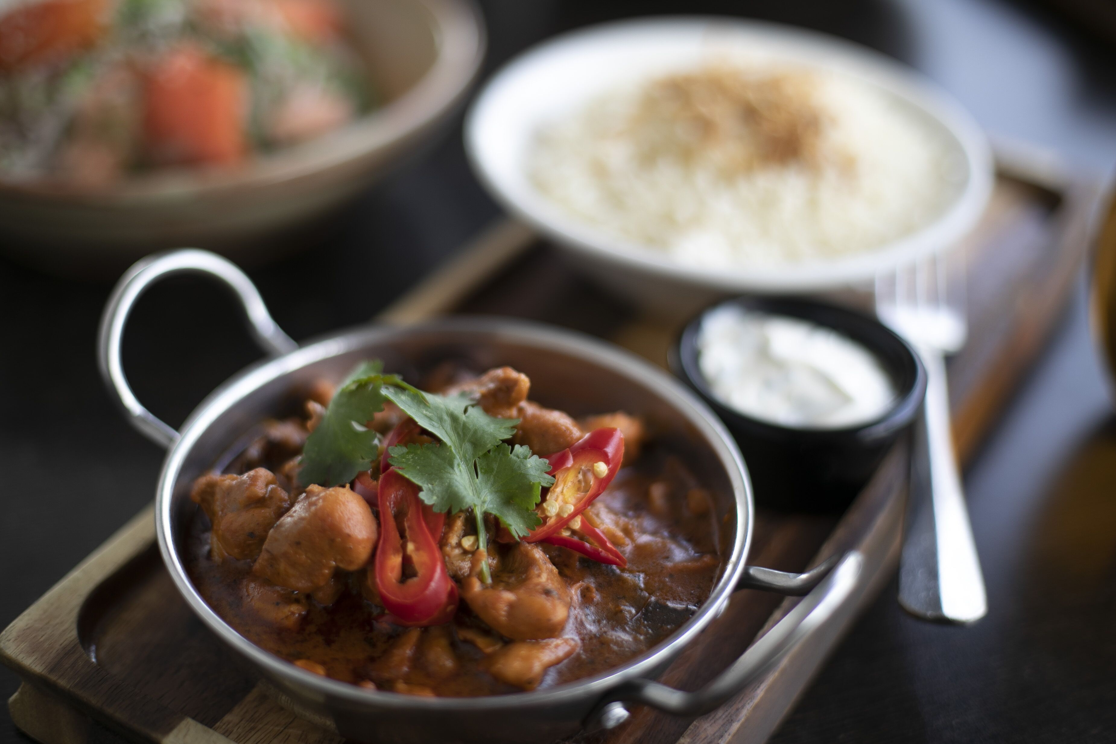 Un tazón de curry con chile rojo y cilantro, un pequeño tazón de salsa blanca y un plato de arroz blanco sobre una tabla de madera.