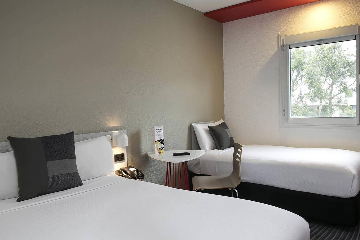Een hotelkamer met twee bedden, een kleine tafel met een afstandsbediening, een telefoon en een raam met uitzicht op bomen.