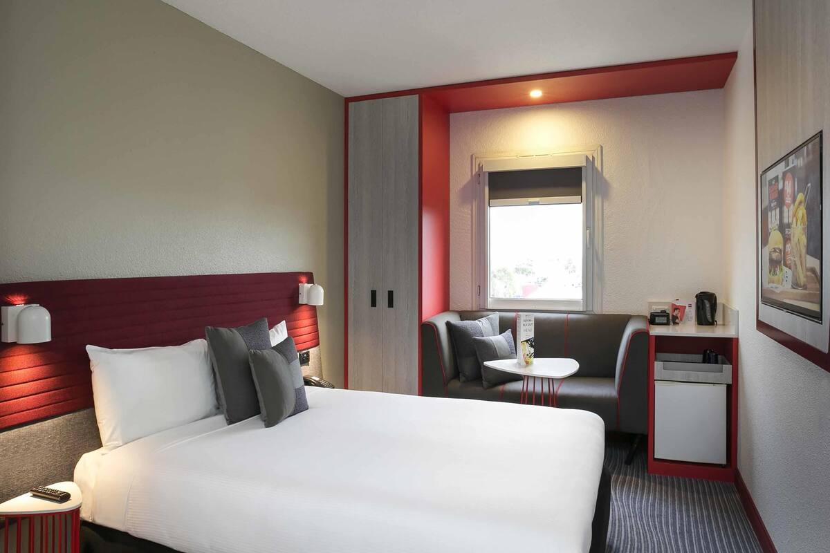 Une chambre d’hôtel avec un lit, un bureau, une chaise, une petite table, une fenêtre et un tableau au mur.