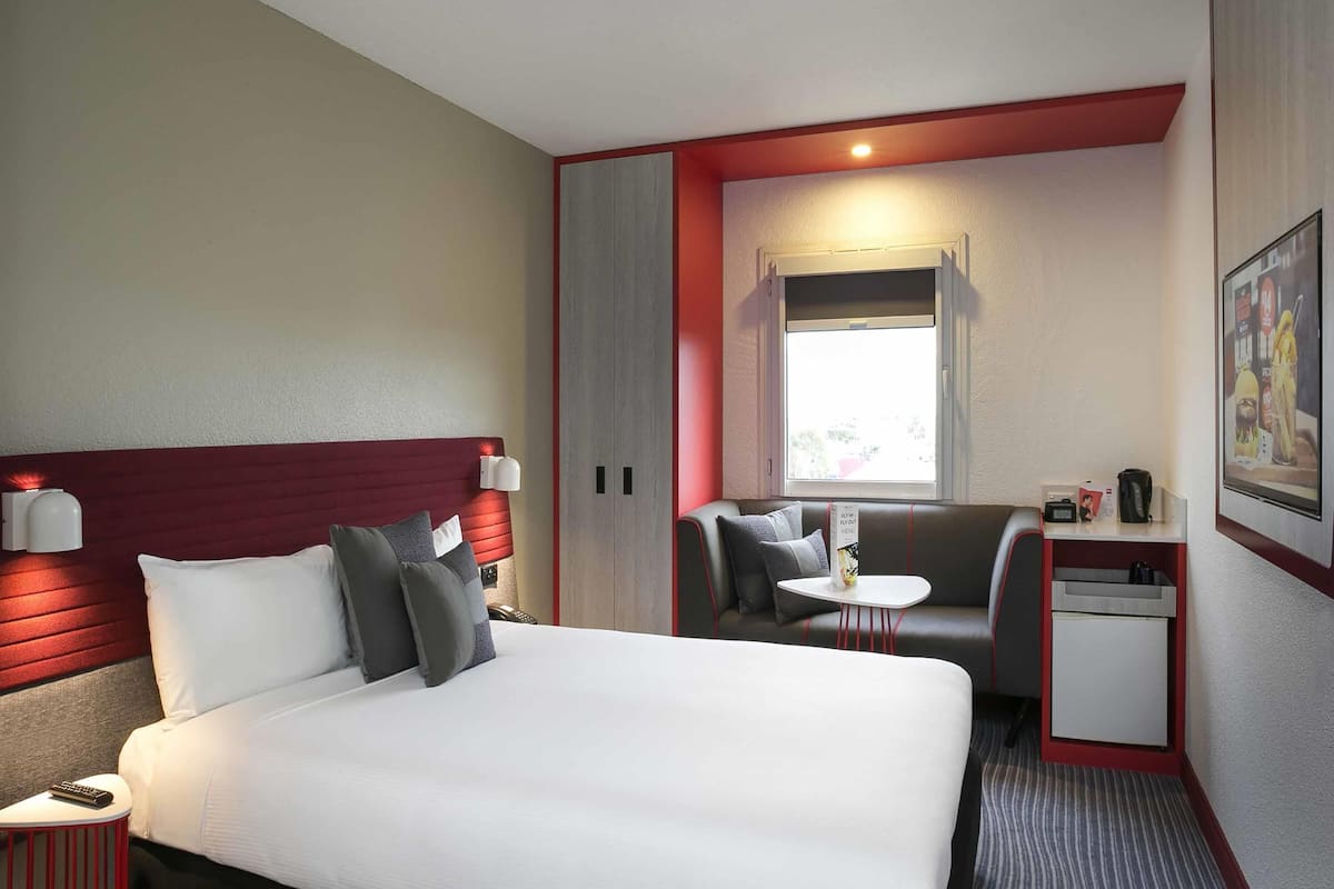 Une chambre dâhĂŽtel avec un lit, un bureau, une chaise, une petite table, une fenĂȘtre et un tableau au mur.