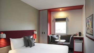 Ein Hotelzimmer mit Bett, Schreibtisch, Stuhl, kleinem Tisch, Fenster und einem Bild an der Wand.