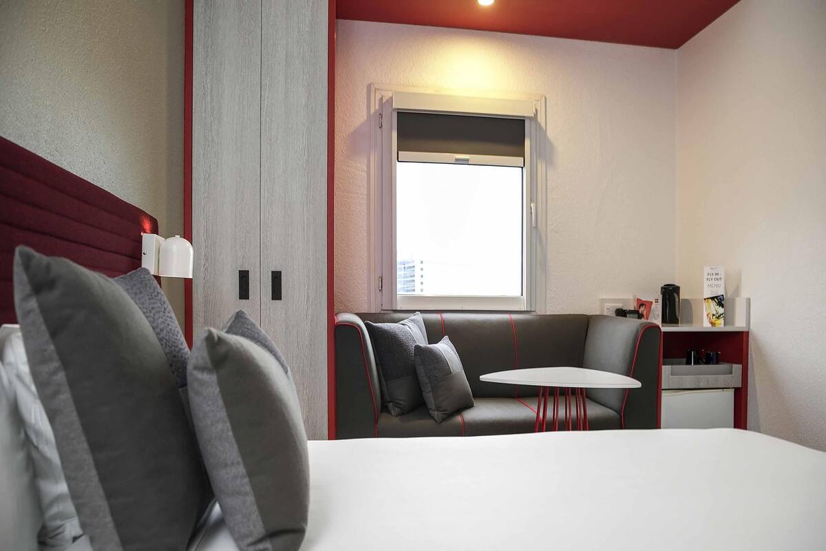 Une chambre d’hôtel comprenant un lit, un canapé, une petite table, une fenêtre avec des stores, une lampe fixée au mur et une étagère avec un menu.