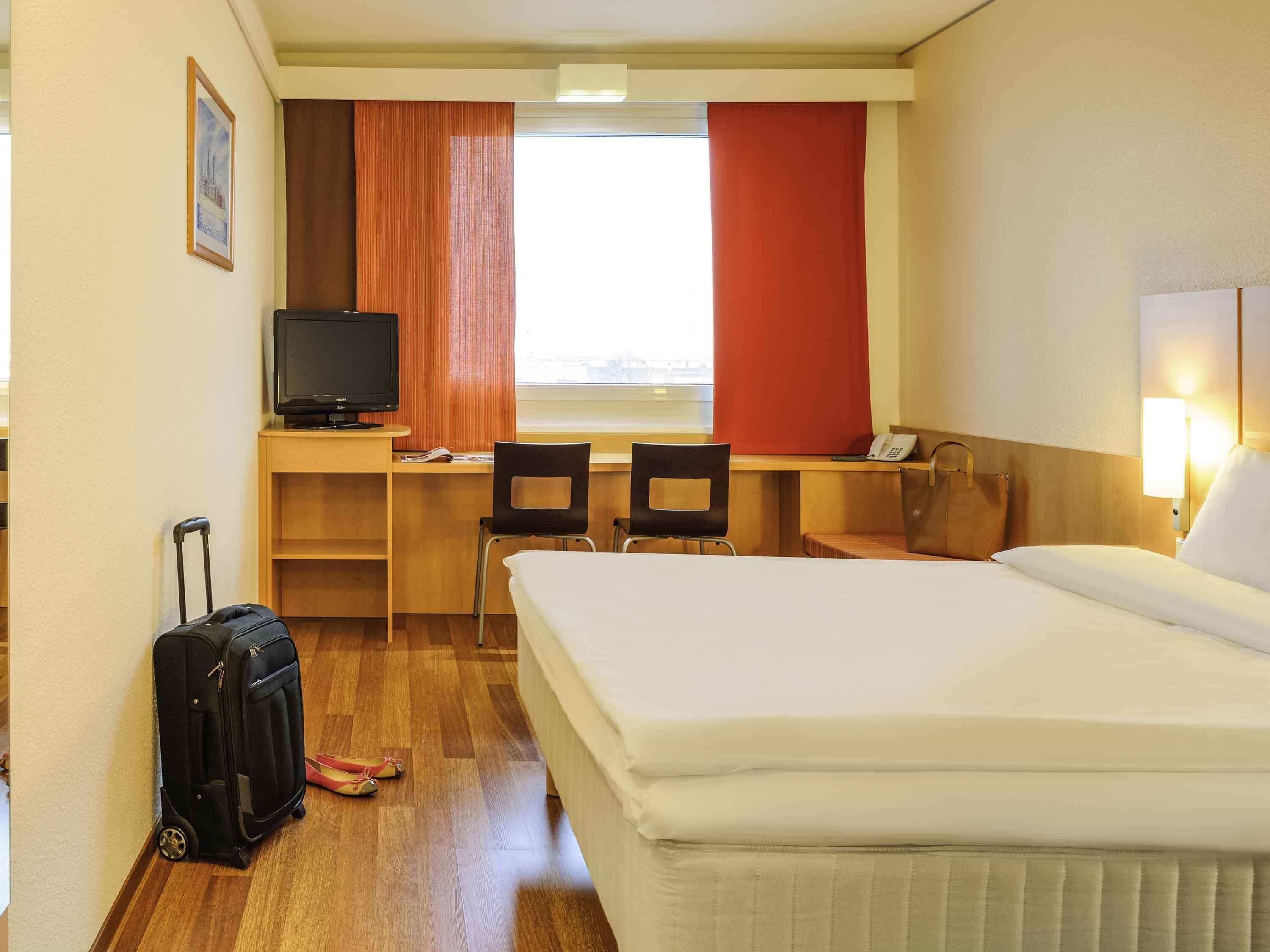 A hotel room with a bed, desk, chairs, a television, a suitcase, and a window with curtains.