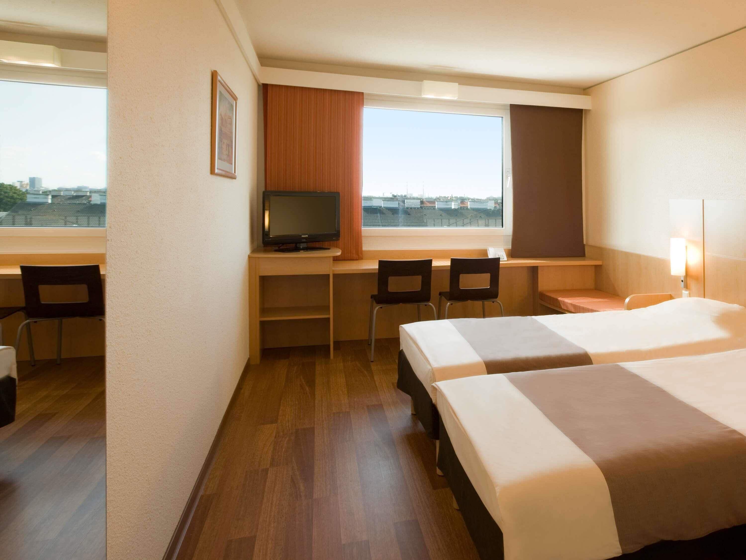 A hotel room with two beds, a desk with a television, and a window with a city view.