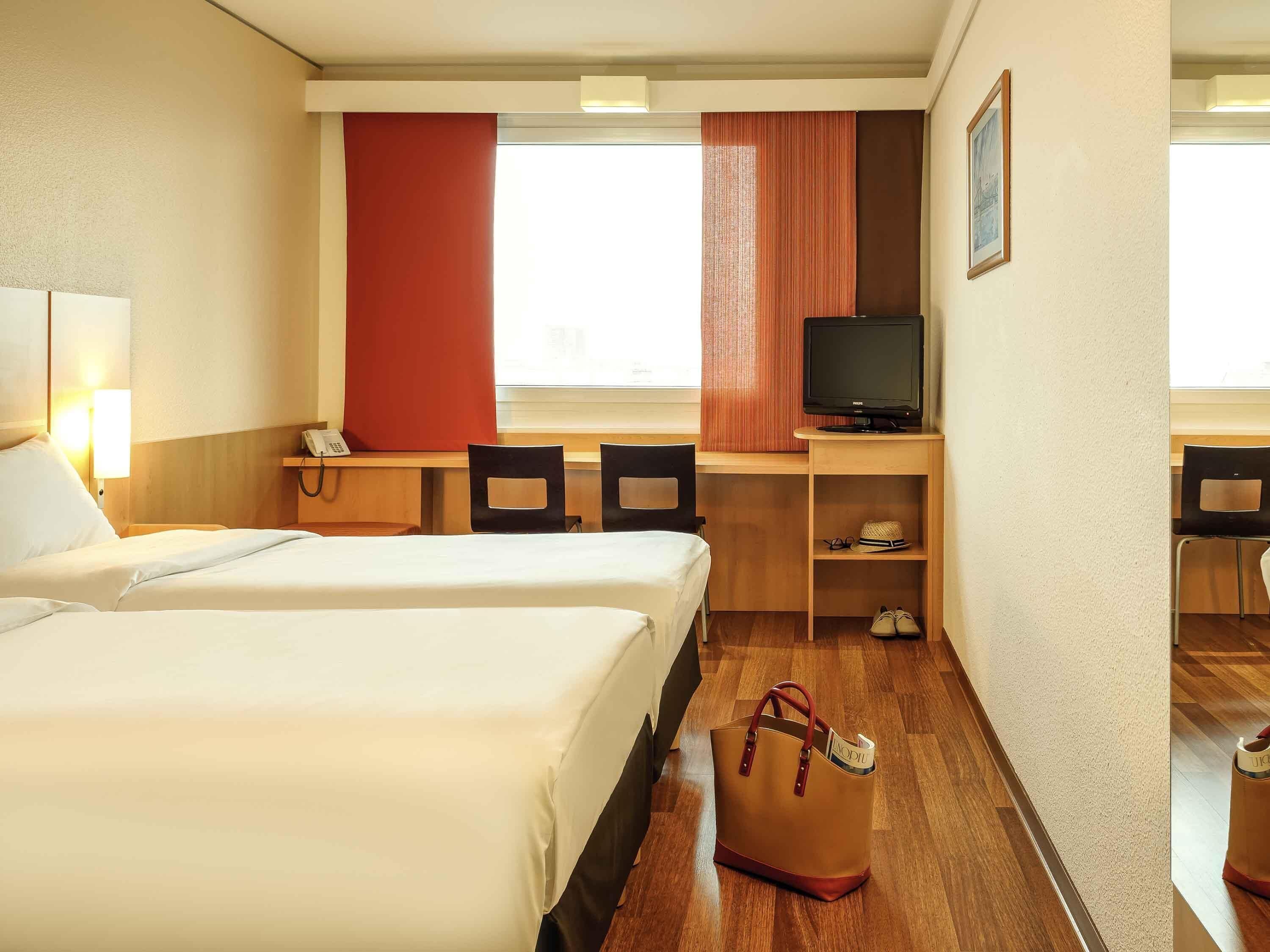 A hotel room with two beds, a desk with a TV, a chair, a phone, and a small shelf with items.