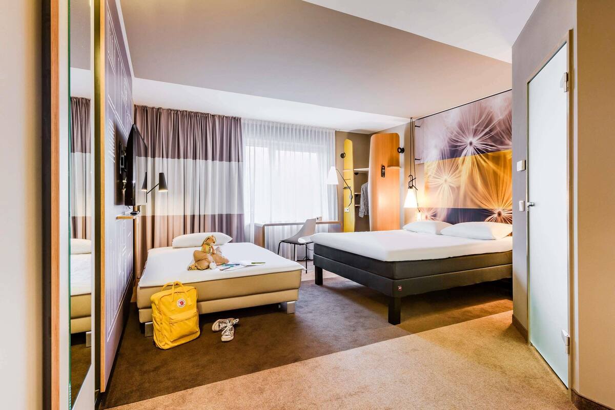 Habitación de hotel con cama, escritorio, silla, armario y un ventanal con cortinas.