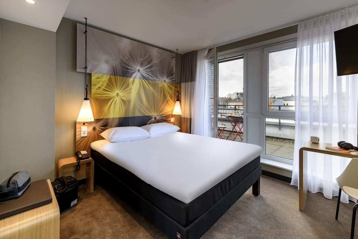 Habitación de hotel con una cama grande, lámparas de noche, un escritorio con silla, televisión y vistas a un paisaje urbano a través de ventanas amplias.