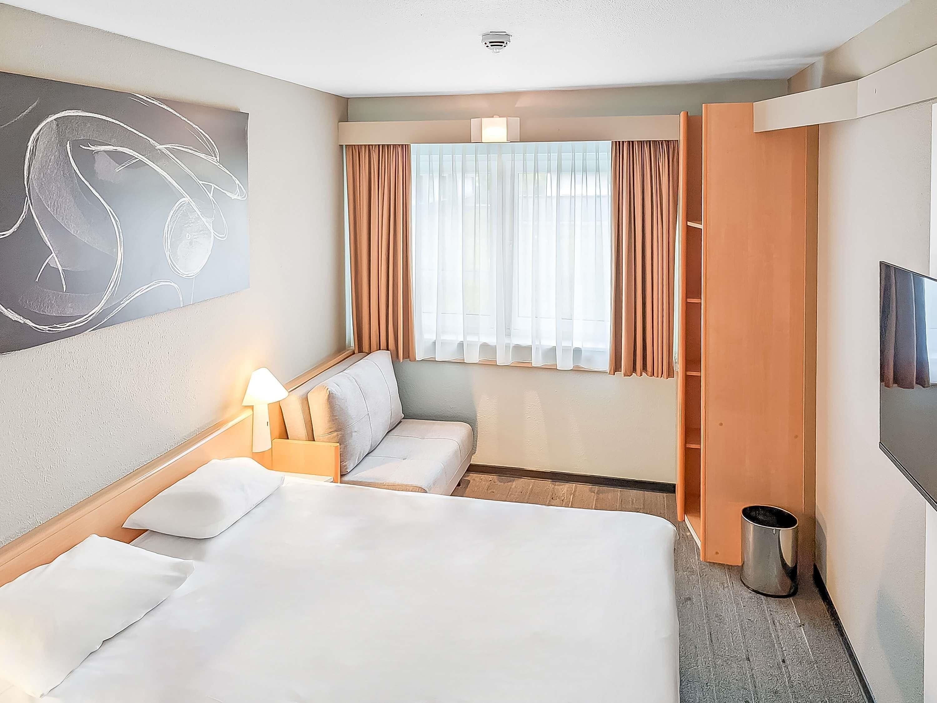 Ein Hotelzimmer mit Bett, Sitzecke, Fernseher, einem Fenster mit Vorhängen und einem Mülleimer.