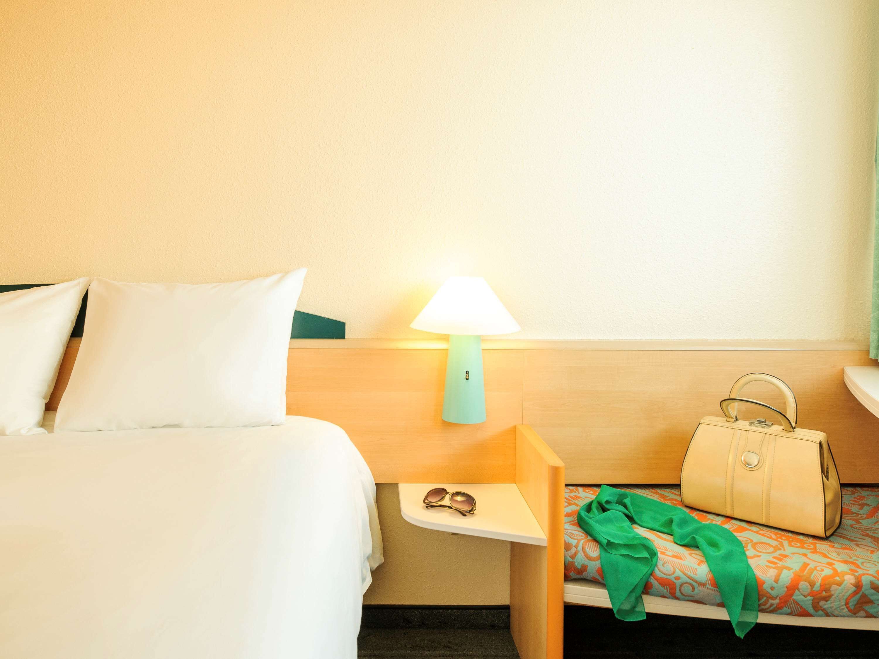 A hotel room with a bed, a nightstand with a lamp, a small table with sunglasses and a handbag, and a patterned chair.