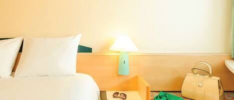A hotel room with a bed, a nightstand with a lamp, a small table with sunglasses and a handbag, and a patterned chair.