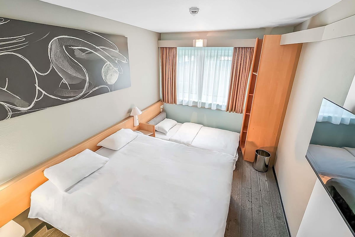 Habitación de hotel con cama, escritorio, silla, lámpara, ventana con cortinas y un cuadro en la pared.