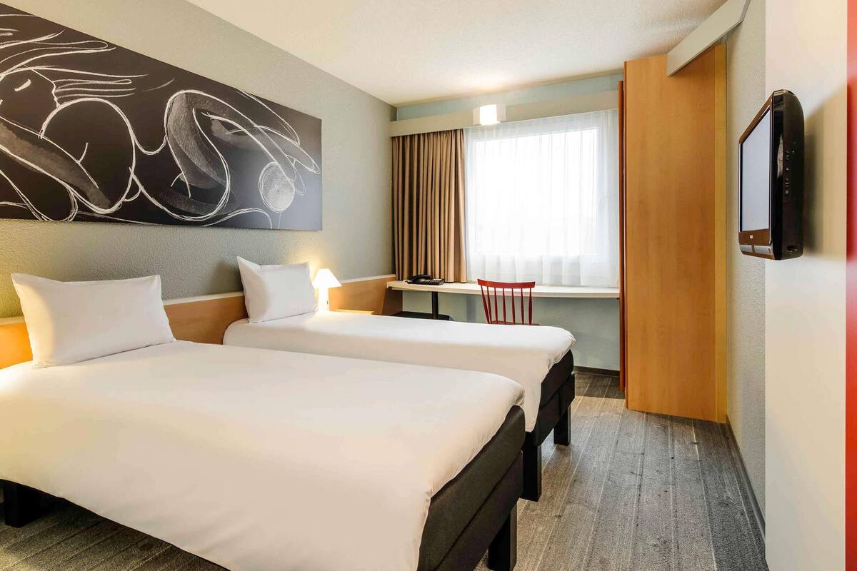 Habitación de hotel con dos camas, un escritorio, una silla, un televisor y un cuadro abstracto grande en la pared.