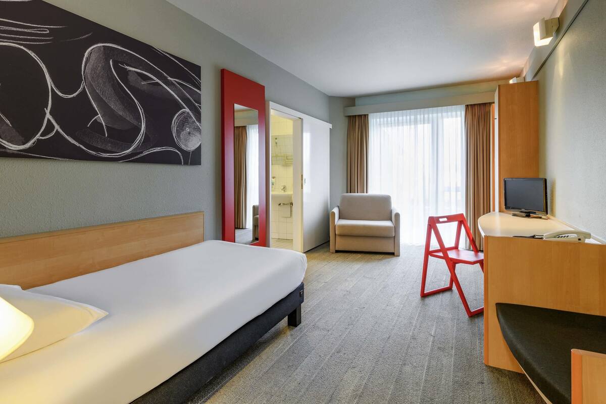 Habitación de hotel con cama, una silla, un escritorio con computadora, televisor, baño visible a través de una puerta abierta y un cuadro abstracto grande en la pared.