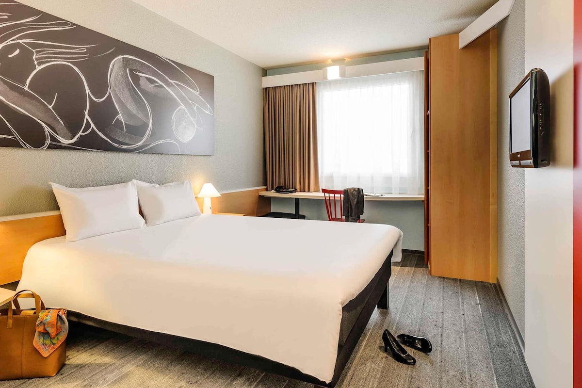 Habitación de hotel con una cama grande, un escritorio, una silla, un televisor y una ventana con cortinas.