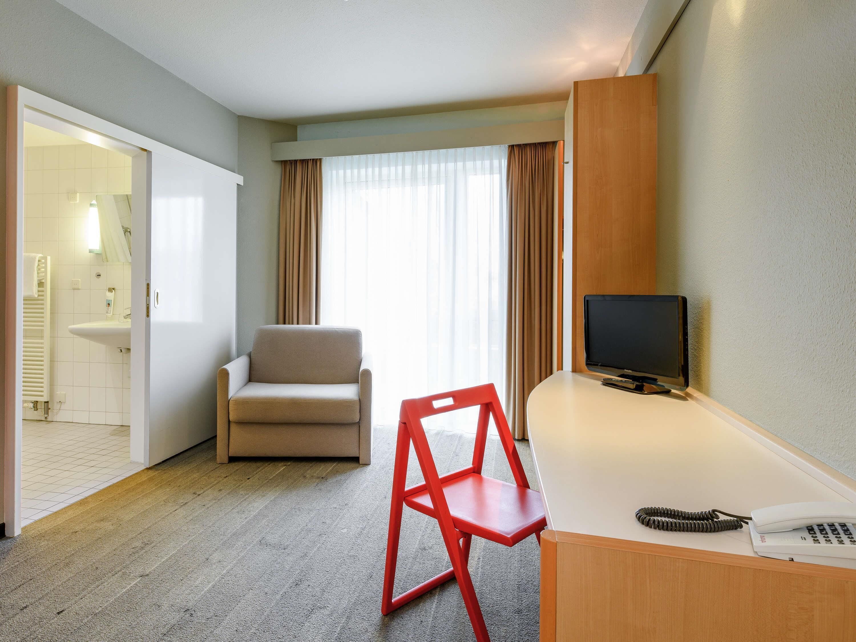 Une chambre d’hôtel avec un lit, une télévision, un bureau, une chaise et une salle de bain visible à travers une porte ouverte.