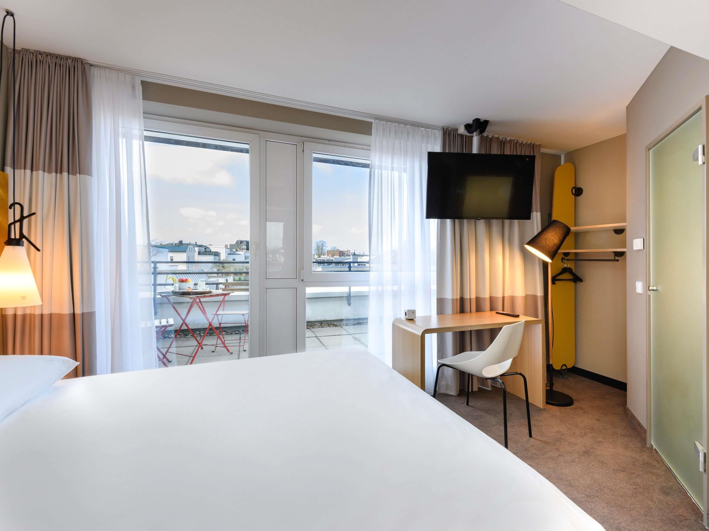 Ein Hotelzimmer mit Bett, Schreibtisch, Stuhl, Fernseher, Lampe und Blick auf die Stadt durch große Fenster.