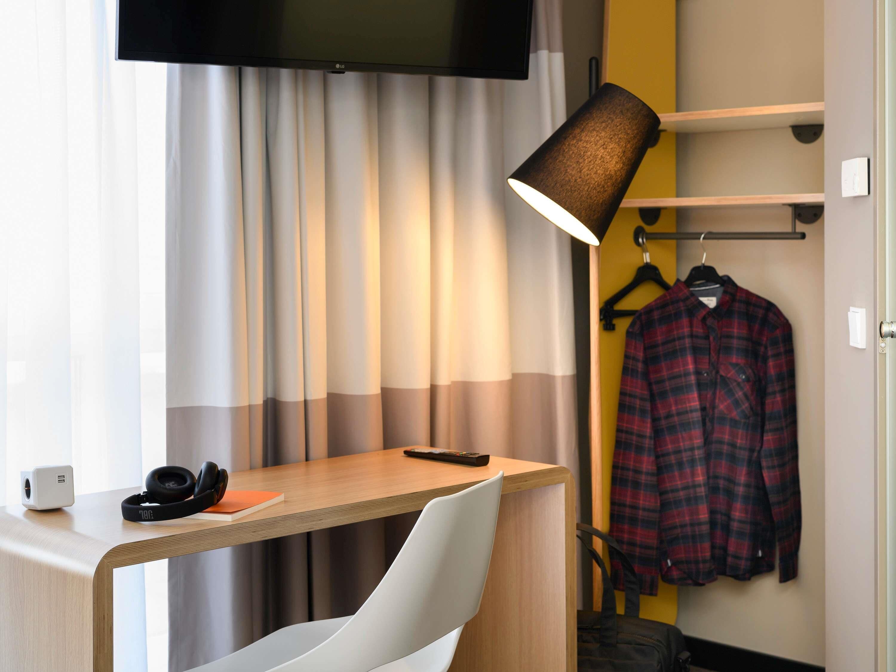 Ein Hotelzimmer mit Schreibtisch, Stuhl, Wandfernseher, Lampe und Kleiderständer mit kariertem Hemd sowie einem kleinen Lautsprecher.