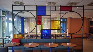 Egy modern étterem, kerek asztalokkal és kék színű, fülkés ülőhelyekkel, geometrikus mintázatú mennyezettel és nagy ablakokkal.