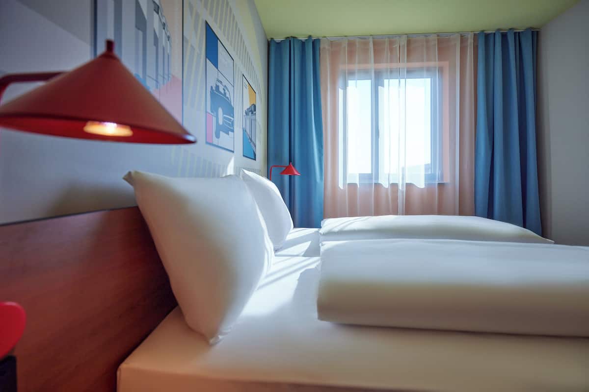A hotel room with a neatly made bed, a red lamp, and a window with blue curtains.