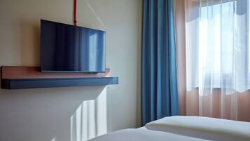 A hotel room with a bed, a mounted TV on a wall, and a window with sheer curtains.