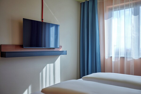 A hotel room with a bed, a mounted TV on a wall, and a window with sheer curtains. - McDreams Hotel Stuttgart-City (Stuttgart)