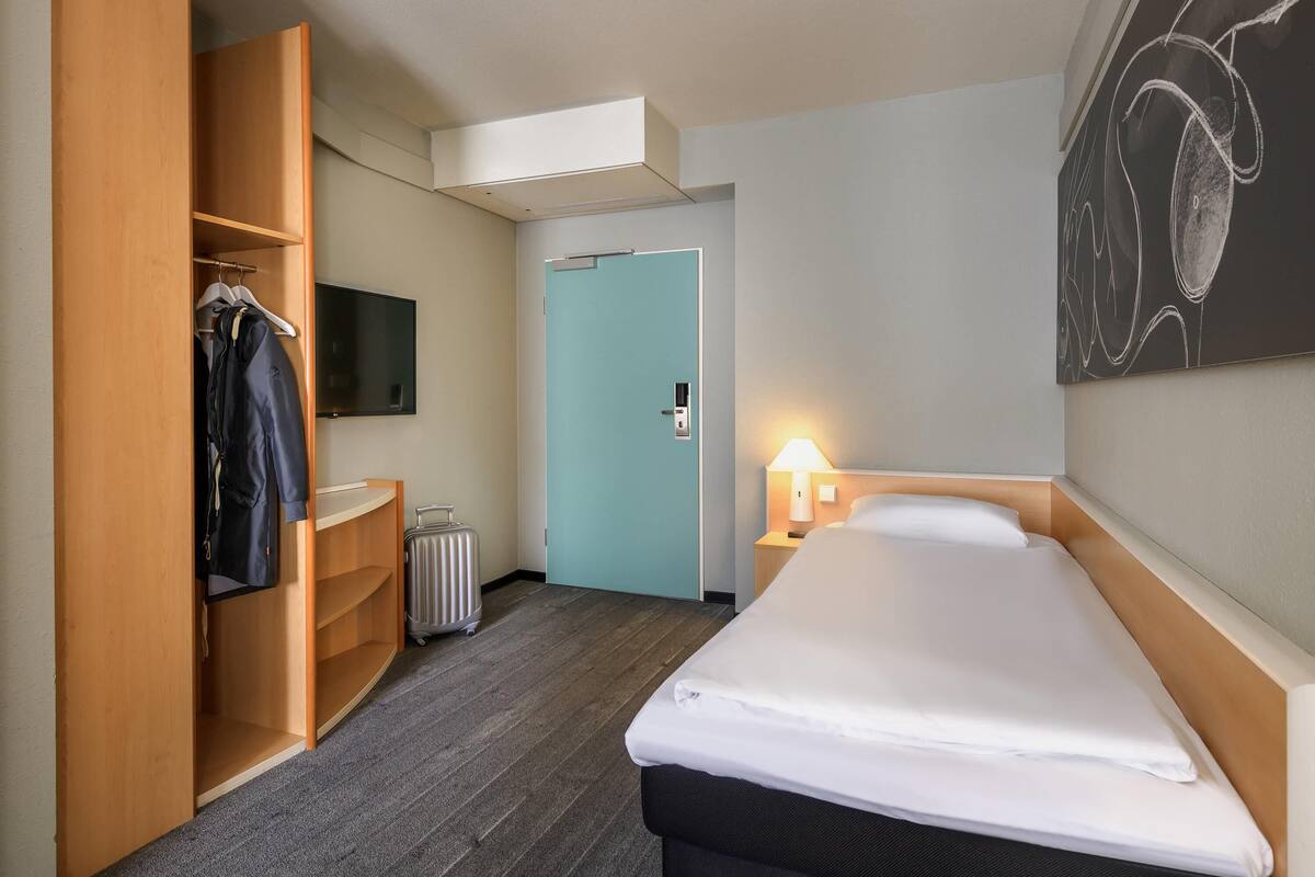 Quarto de hotel com cama, guarda-roupa, televisão, porta e uma obra de arte pendurada na parede.