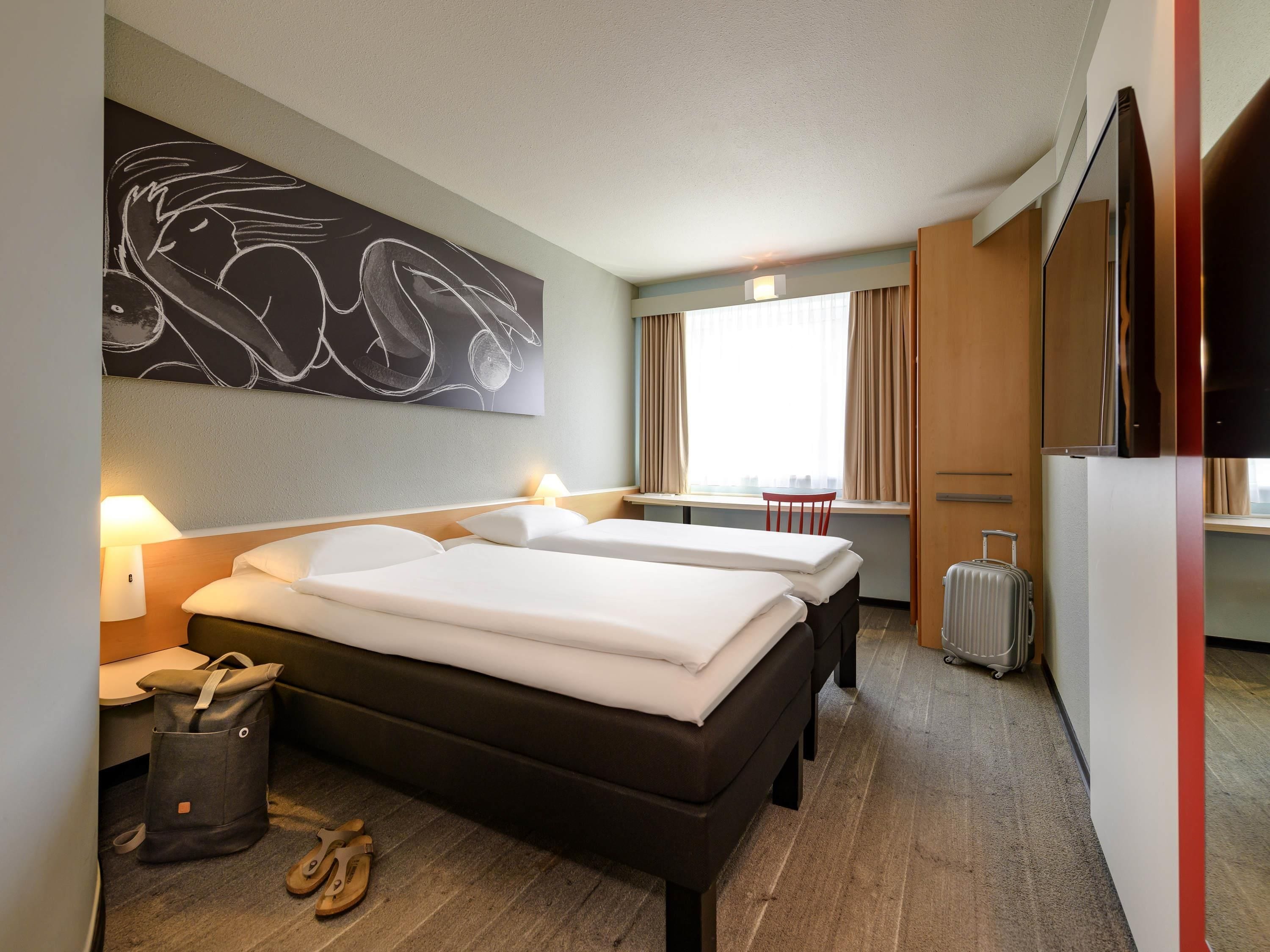 A hotel room with two beds, a desk, a chair, a suitcase, and a large abstract painting on the wall.