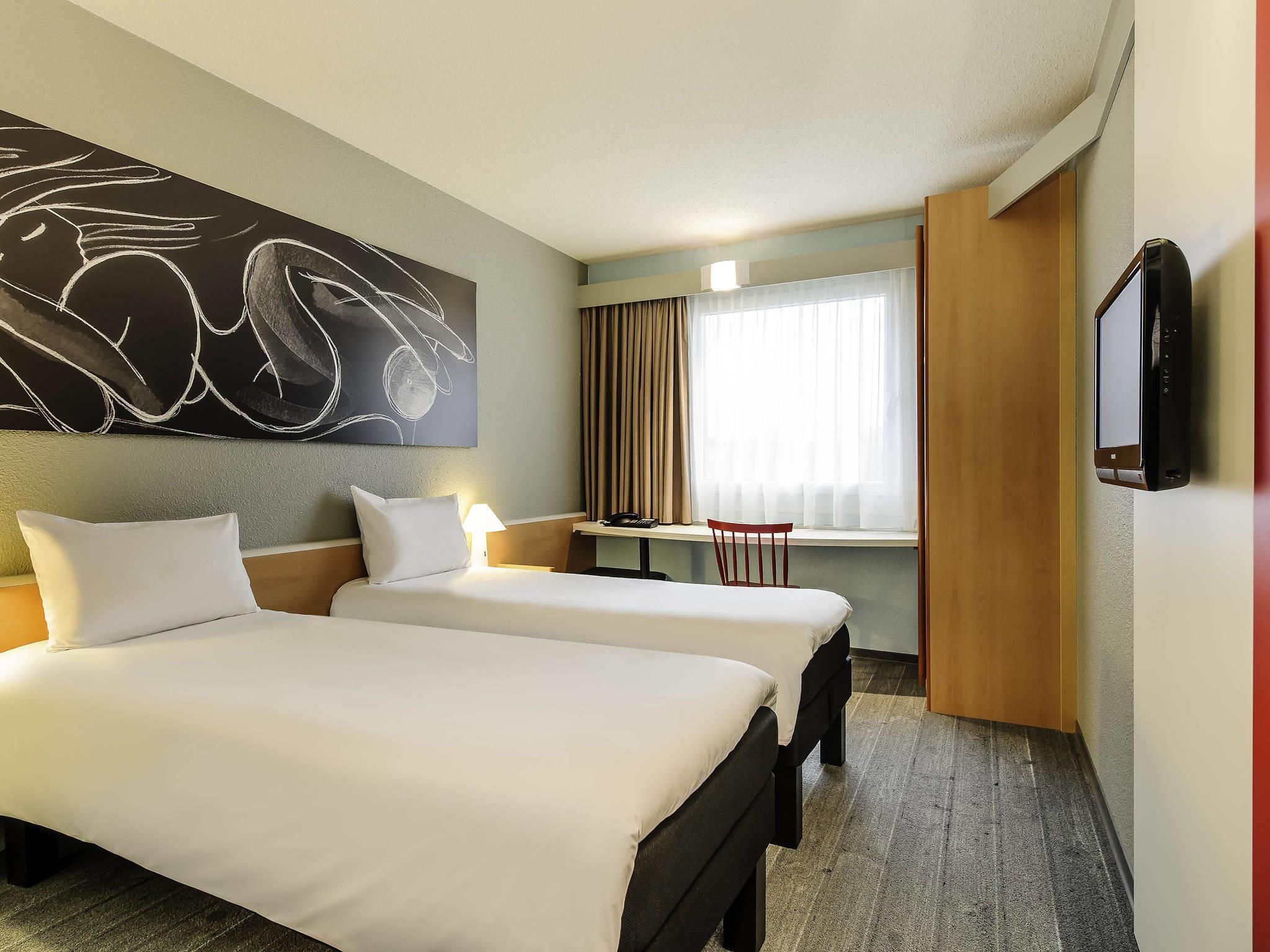 A hotel room with two beds, a desk, a chair, a TV, and a large abstract painting on the wall.