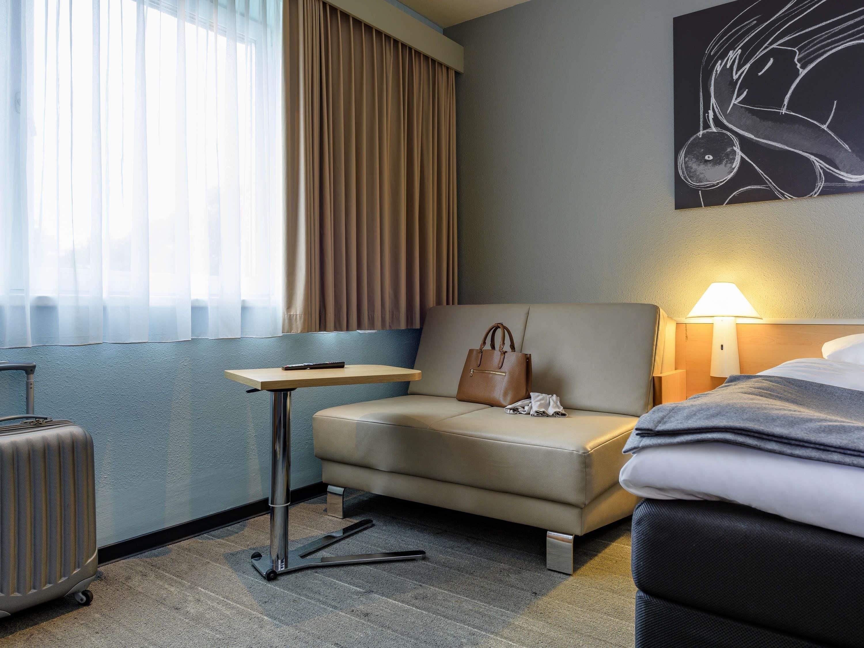 A hotel room with a bed, a sofa, a table with a remote control, a suitcase, and a lamp.