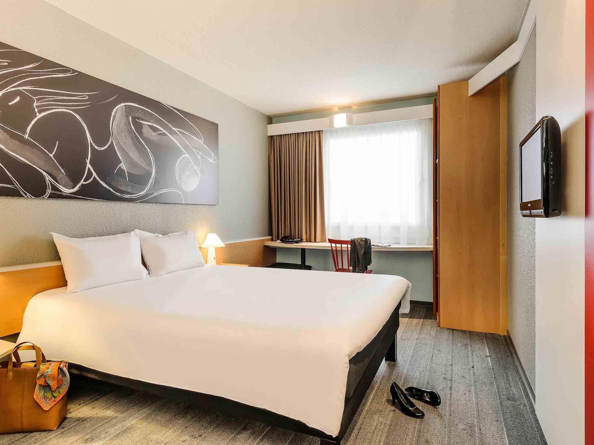 A hotel room with a large bed, a desk, a chair, a TV, and a window with curtains.