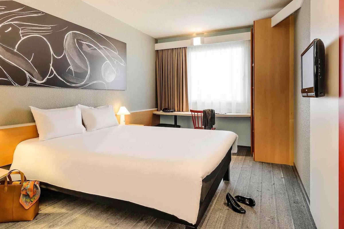Quarto de hotel com uma cama grande, uma mesa, uma cadeira, uma TV e uma janela com cortinas.