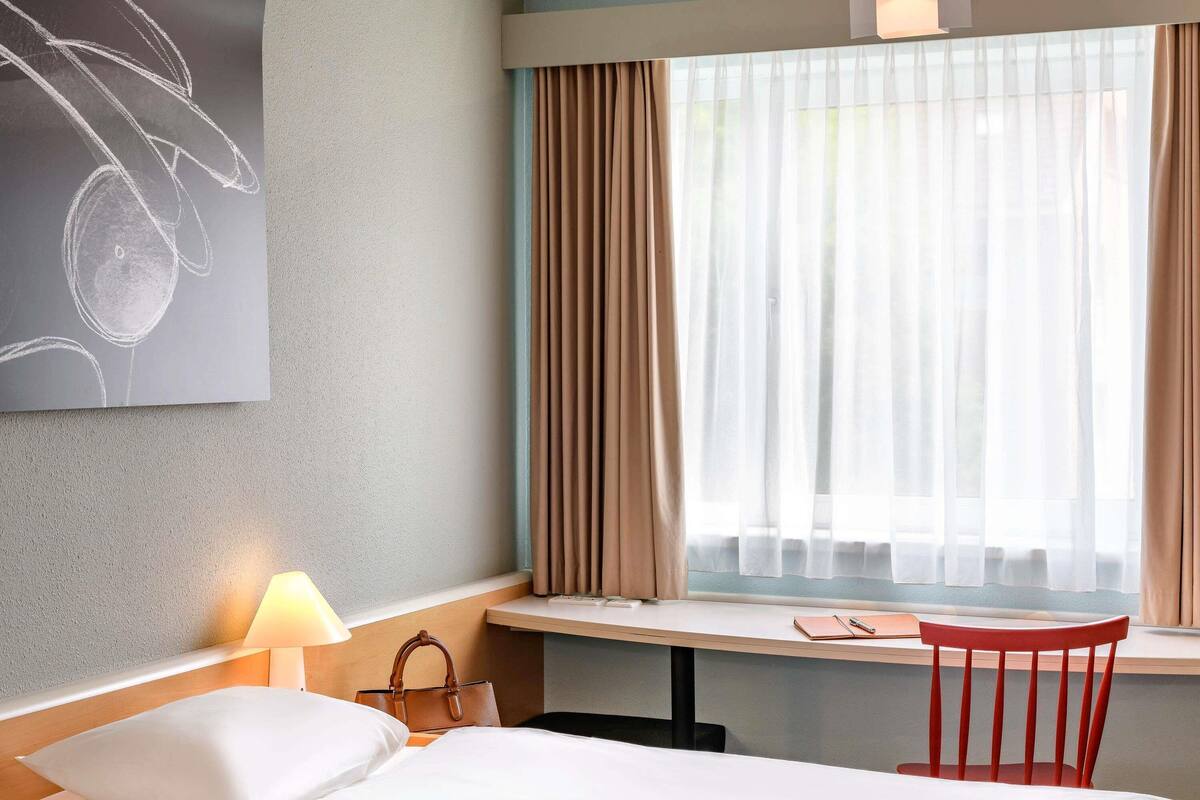 Quarto de hotel com cama, escrivaninha, cadeira, luminária e janela com cortinas.
