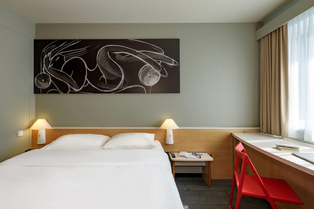 A hotel room with a bed, desk, and chair, a painting on the wall, and a window with curtains.