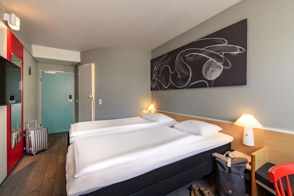 Quarto de hotel com cama, duas luminárias de cabeceira, uma obra de arte na parede, um guarda-roupa vermelho, uma mala e uma cadeira.