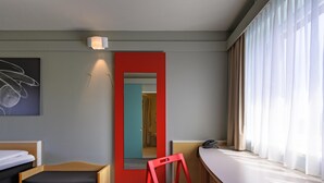 Ein Hotelzimmer mit einem Bett, einem roten Sessel, einem Schreibtisch, einem Fenster mit Vorhängen, einem Bild an der Wand und einer Tür zu einem anderen Raum.