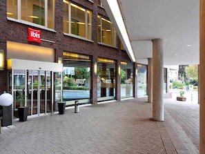 Entrada de hotel con fachada de vidrio, un letrero rojo de 'ibis' y un letrero de 'Hopfen Malz'.
