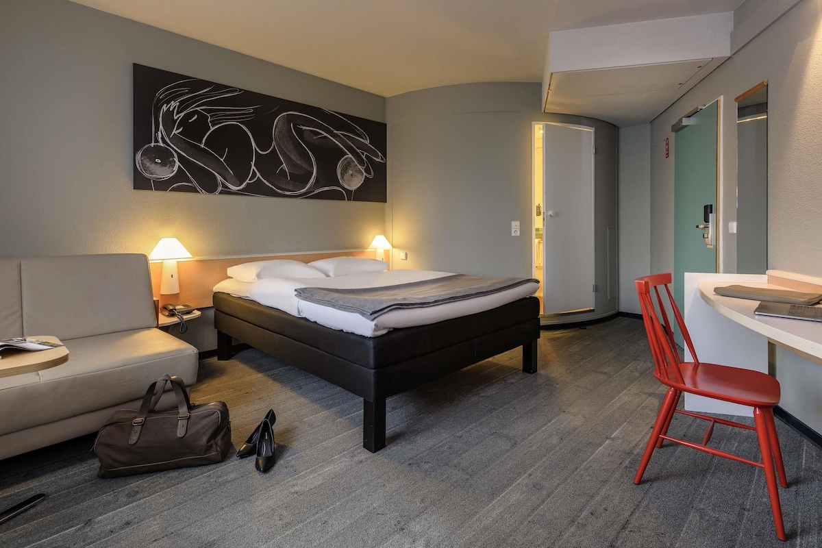 A hotel room with a bed, bedside lamps, a desk with a chair, a coat rack, and a door.