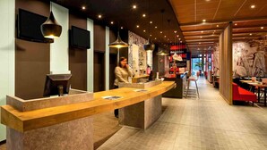 A modern hotel reception area with a curved reception desk, a TV mounted on the wall, and a person standing behind the counter. - ibis Hamburg Alsterring (Hamburg)