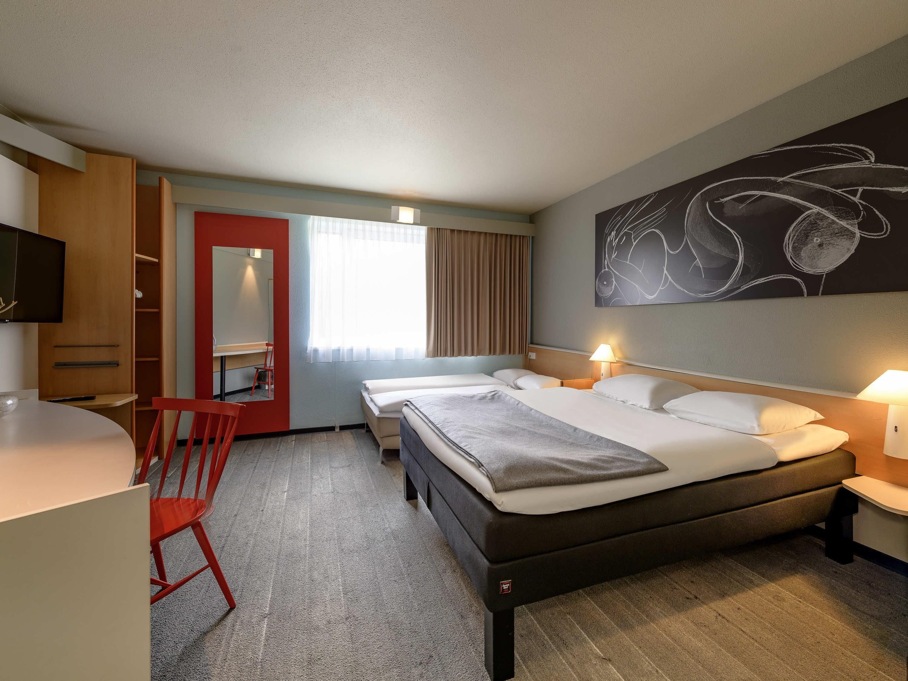 Une chambre d’hôtel comprenant un lit, des tables de chevet, un bureau, une chaise, un téléviseur et un grand tableau abstrait accroché au mur.