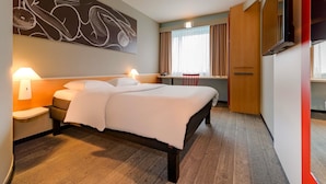 A hotel room with a bed, bedside lamps, a desk, a chair, a TV, and a large abstract painting on the wall. - ibis Hamburg Alsterring (Hamburg)