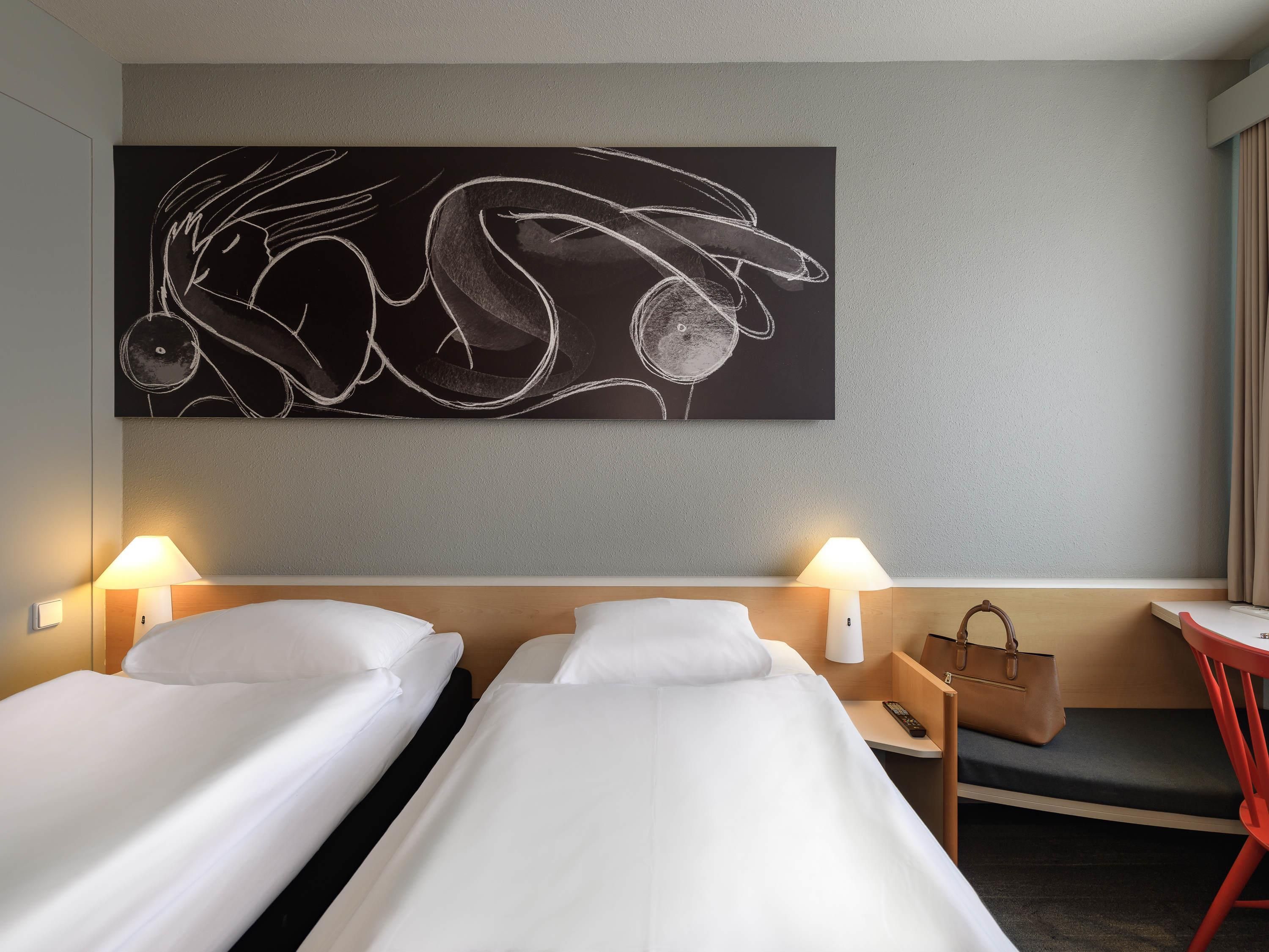 A hotel room with two single beds, a large abstract artwork on the wall, bedside lamps, a small table with a remote control, and a handbag on the nightstand.