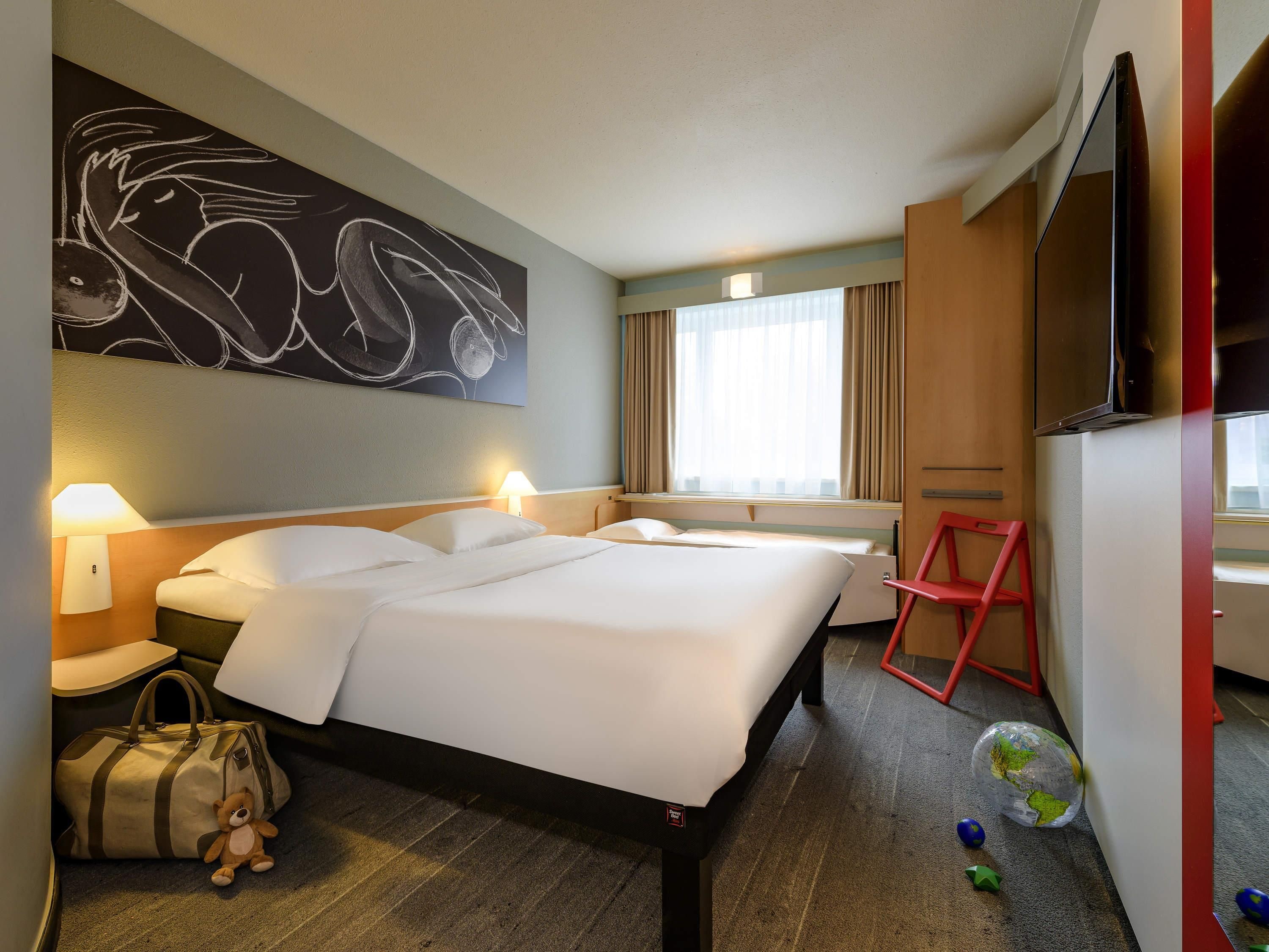 A hotel room with a bed, bedside lamps, a large abstract painting, a television, a window with curtains, and a red chair.