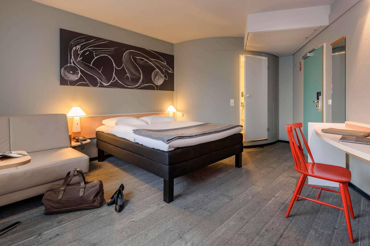 Quarto de hotel com cama, abajures, escrivaninha, cadeira, bolsa e um par de sapatos.