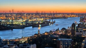 A waterfront cityscape at dusk with illuminated buildings and cranes, a large ship docked, and a river. - ibis Hamburg Alsterring (Hamburg)