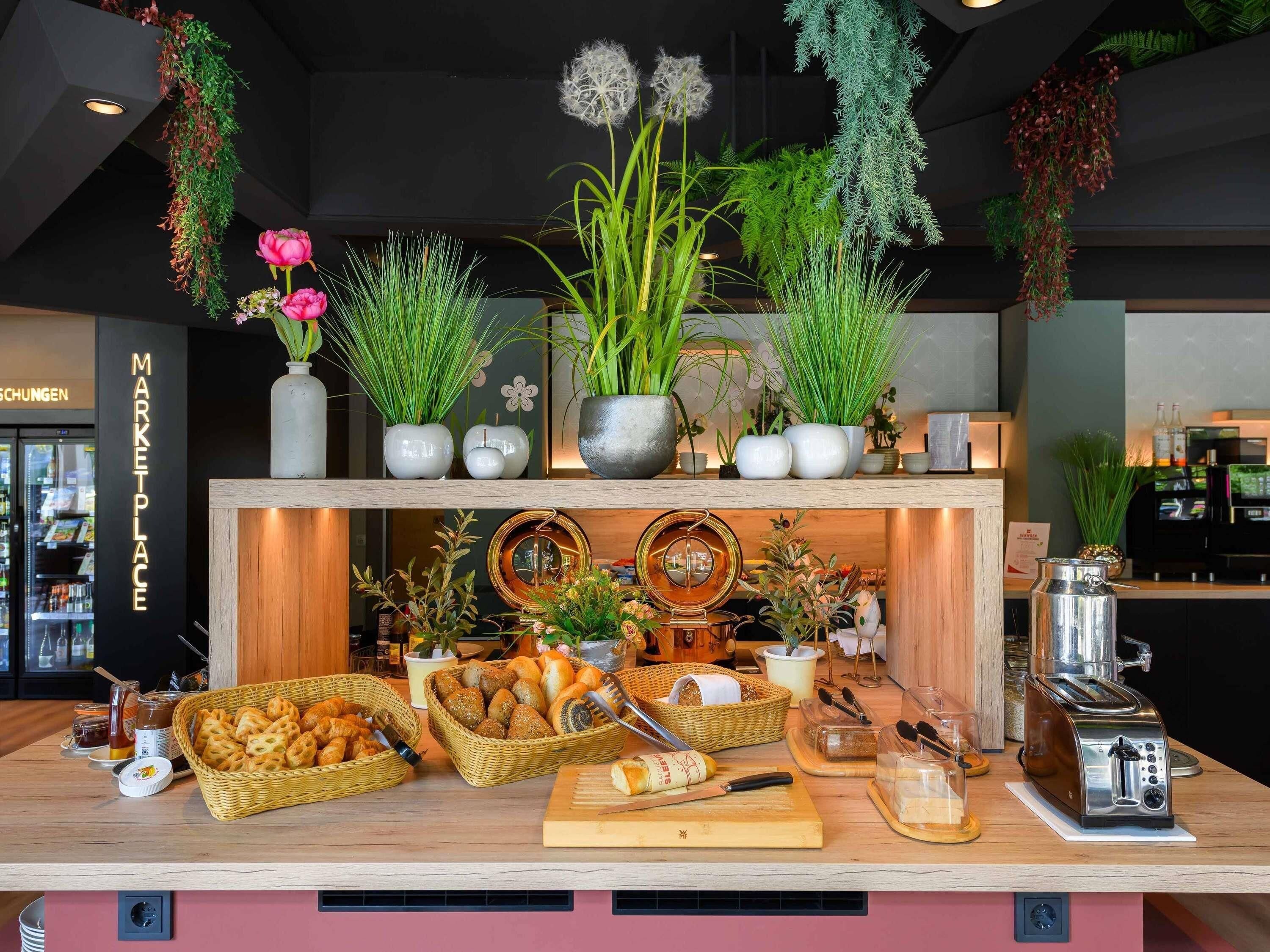 Mostrador de panadería con diversos productos horneados, una máquina de café y plantas decorativas.