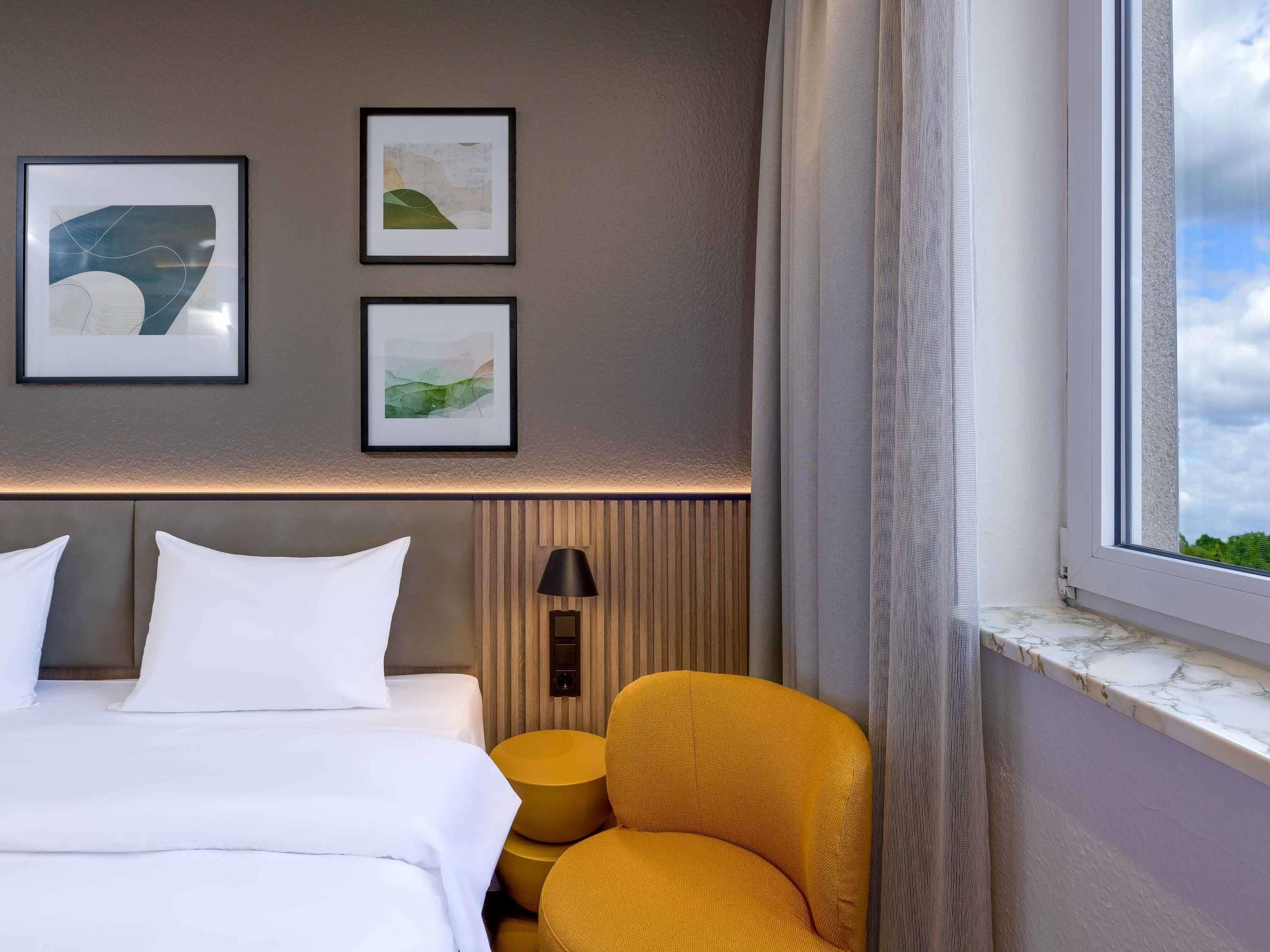 A hotel room with a bed, two framed artworks, a lamp, a yellow chair, and a window with a view.