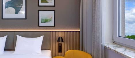 A hotel room with a bed, two framed artworks, a lamp, a yellow chair, and a window with a view.