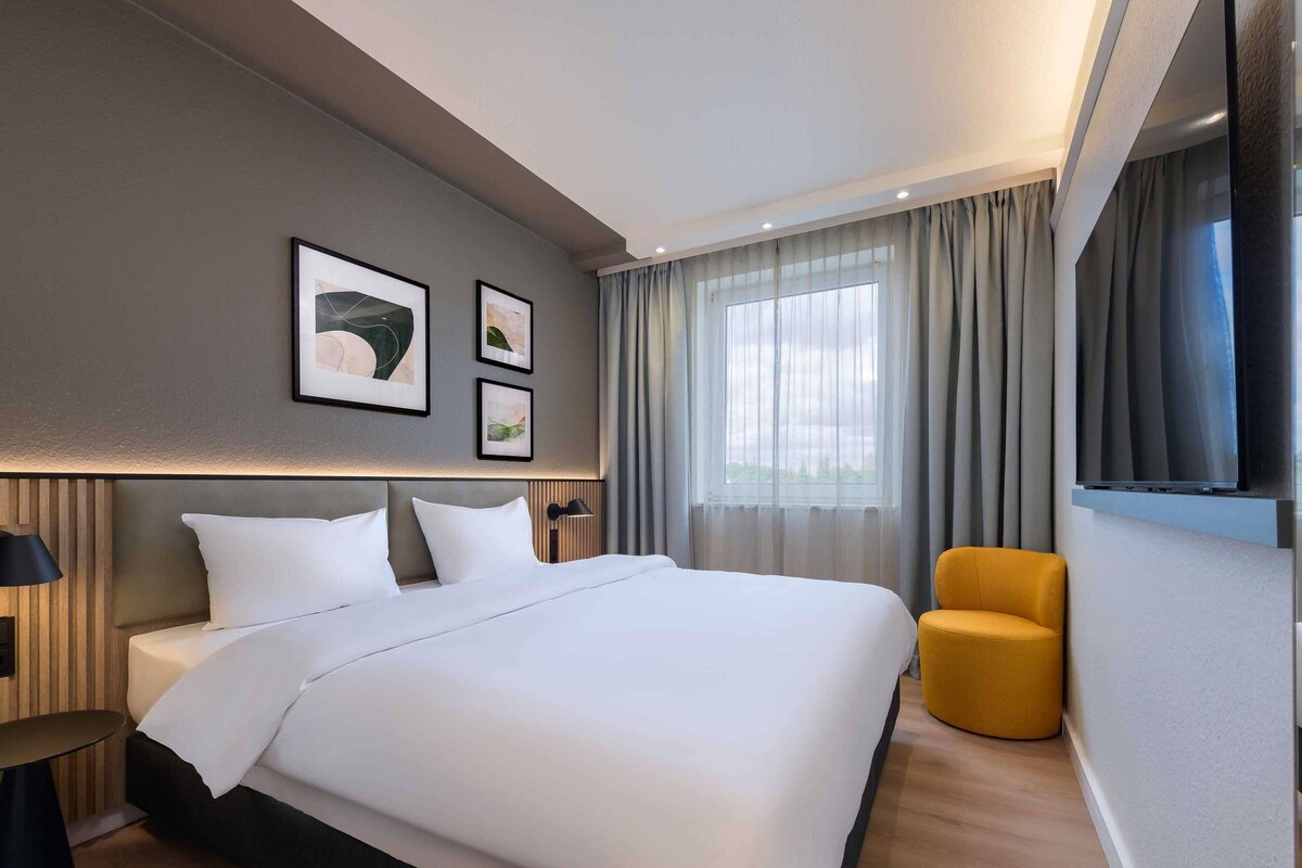 Una habitación de hotel moderna con una cama grande, un sofá, un televisor y un ventanal con cortinas.